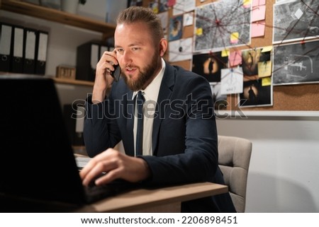 Handsome police officer sitting beside computer, talking on a cell phone, evidence board on background, copy space