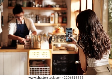 Young woman filming coffeeshop worker packing orders in paper bags Royalty-Free Stock Photo #2206898533