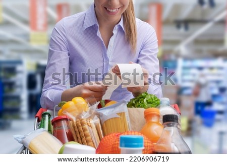Smiling woman leaning on the shopping cart and checking the grocery receipt Royalty-Free Stock Photo #2206906719