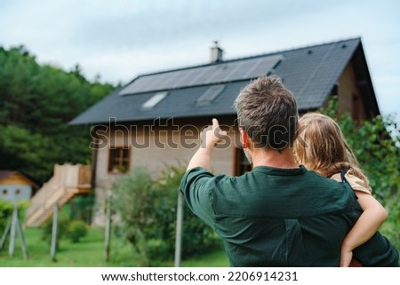 Rear view of dad holding her little girl in arms and showing at their house with installed solar panels. Alternative energy, saving resources and sustainable lifestyle concept. Royalty-Free Stock Photo #2206914231