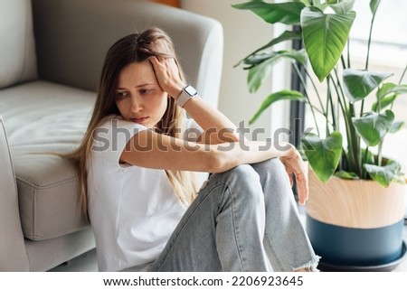 Unhappy young caucasian woman with blonde hair thinking about bad relationships problems, break up with boyfriend. Worried millennial girl sitting on floor in bedroom near chair and green plant alone Royalty-Free Stock Photo #2206923645