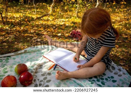 a little red-haired girl draws on a picnic in autumn on a blanket. there are apples nearby
