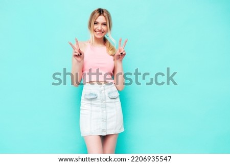 Young beautiful smiling blond female in trendy summer jeans skirt. carefree woman posing near blue wall in studio. Positive model having fun indoors. Cheerful and happy. Shows peace sign