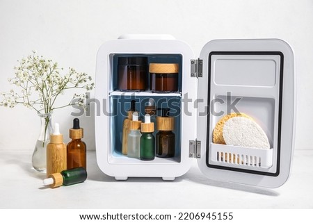 Small refrigerator with natural cosmetics and flowers in vase on white background