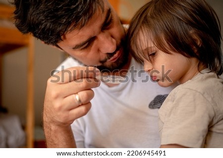 A sick child is sitting on his father's lap. Dad tries to get his son drunk with medicine Royalty-Free Stock Photo #2206945491