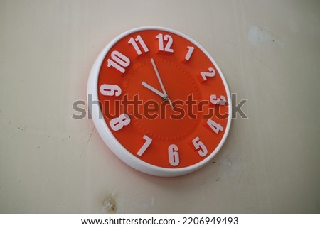 A photo of a round orange wall clock with hands indicating 10 am
