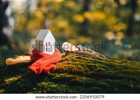 Miniature house in a red scarf on an autumn background with moss and yellow leaves. The concept of passive house heating. Thermal insulation of a building or dwelling. Energy crisis. Royalty-Free Stock Photo #2206950079