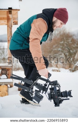 Videographer man walking on snow, shooting footage, using camera mounted on gimbal stabilizer equipment in winter.