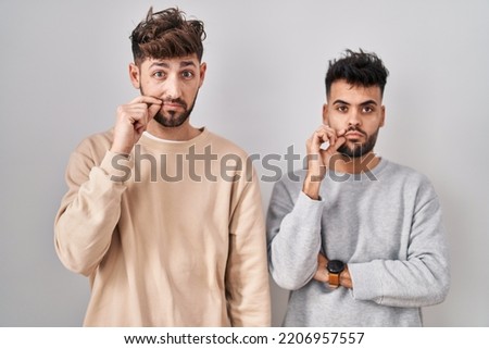 Young homosexual couple standing over white background mouth and lips shut as zip with fingers. secret and silent, taboo talking 