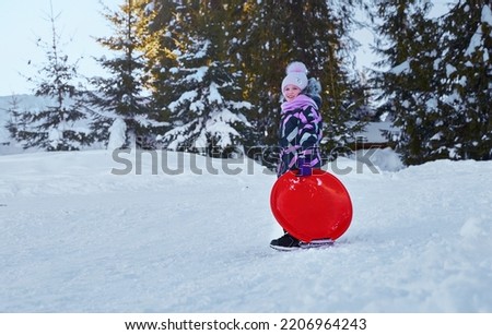 Little girl enjoying the winter sledding time. Child playing and having fun riding on a snowy hill. Slide down from snow slope sitting in plate