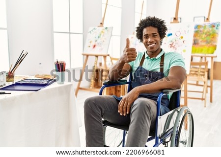 Young african american artist man sitting on wheelchair at art studio doing happy thumbs up gesture with hand. approving expression looking at the camera showing success. 