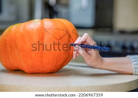 A woman draws a scary face on an orange pumpkin with a black marker in the kitchen. Halloween holiday design concept.