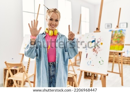 Young caucasian girl at art studio showing and pointing up with fingers number six while smiling confident and happy. 