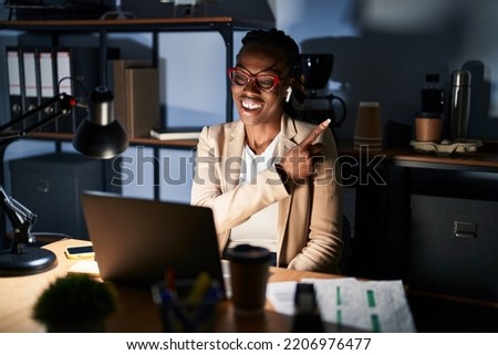 Beautiful black woman working at the office at night cheerful with a smile of face pointing with hand and finger up to the side with happy and natural expression on face 