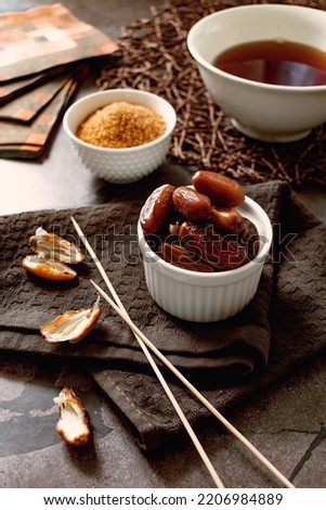 Sweet dried dates in glucose syrup, in a small plate, next to long skewers, brown sugar and tea, dried fruit dessert for sweet tooth, healthy tasty food