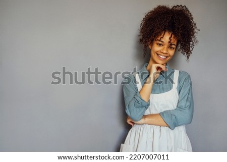 Young black woman wearing beige apron isolated on grey background with copy space. Portrait of successful african american woman on gray wall. Smiling black waitress looking at camera.