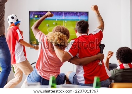 Group of young friends having fun watching football match on TV, drinking beer and cheering; football fans watching game at home celebrating after their team scoring a goal. Focus on the man in jersey Royalty-Free Stock Photo #2207011967