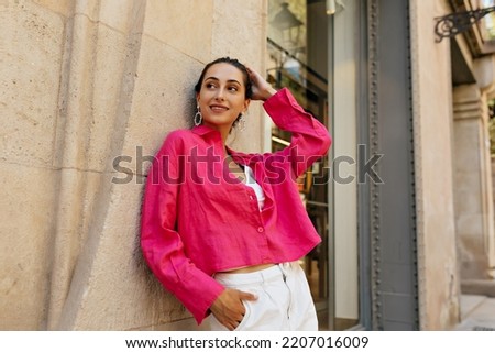 Slender stylish girl modestly poses for photo against beige wall. Smiling lady in summer pink blouse with dark hair is having good on warm day. Vacation pastime concept