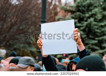 A guy with a white paper banner in their hands protests on the street. Young. Outdoors. Protester. Protestor. Anger. Back. Community. Crisis. Equality. Fight. Fighting. Fist. March. Politics
