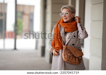 Smiling Woman Outdoor Portrait. Short blonde hair fashion model wears stylish clothes, double-breasted jacket, leather handbag, ochre knitted scarf and glasses. Fashion trend of autumn or spring Royalty-Free Stock Photo #2207045719