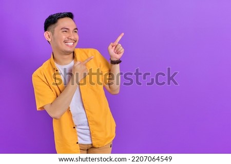Cheerful handsome young Asian man pointing two fingers aside at copy space isolated on purple background Royalty-Free Stock Photo #2207064549