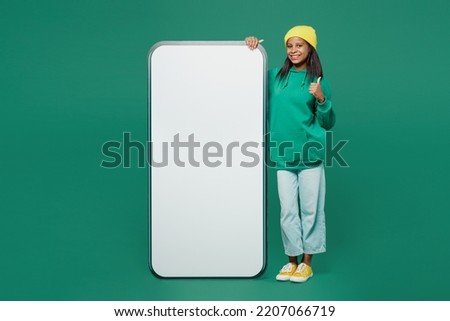 Full body little kid teen girl of African American ethnicity 13-14 years old wear casual hoody hat big blank screen mobile cell phone with area show thumb up isolated on plain dark green background