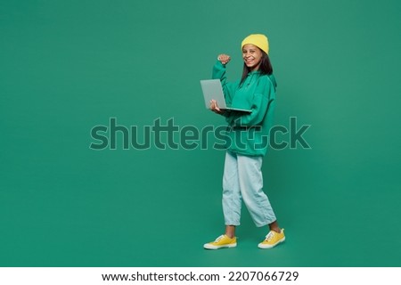 Full body fun IT little kid teen girl of African American ethnicity 13-14 years old wear casual hoody hat hold use work on laptop pc computer do winner gesture isolated on plain dark green background Royalty-Free Stock Photo #2207066729