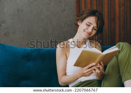 Young smart happy woman wears white tank shirt read book novel sit on blue sofa couch stay at home hotel flat rest relax spend free spare time in living room indoors grey wall. People lounge concept