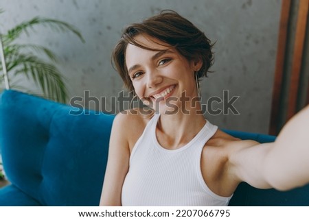Young woman wear white tank shirt doing selfie shot pov on mobile cell phone sit on blue sofa couch stay at home hotel flat rest relax spend free spare time in living room indoor People lounge concept