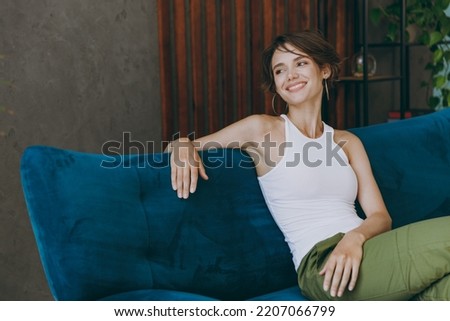 Young minded happy woman 20s wears white tank shirt look aside sit on blue sofa couch stay at home hotel flat rest relax spend free spare time in living room indoors grey wall. People lounge concept Royalty-Free Stock Photo #2207066799