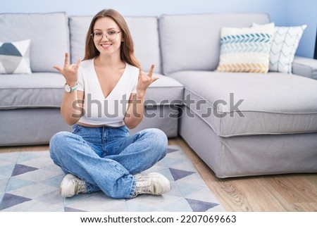 Young caucasian woman sitting on the floor at the living room shouting with crazy expression doing rock symbol with hands up. music star. heavy music concept. 