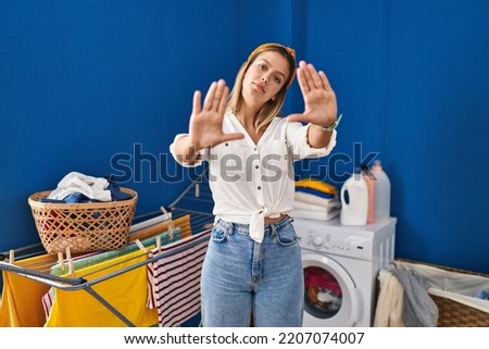Young blonde woman at laundry room doing frame using hands palms and fingers, camera perspective 