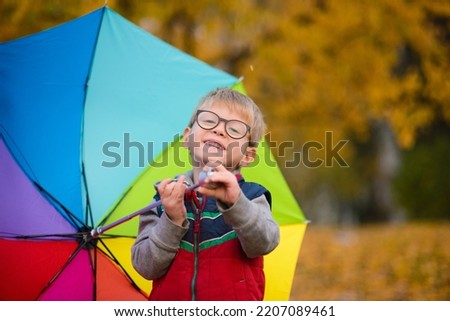 portrait of a child boy in glasses, standing under a rainbow umbrella, against the background of autumn leaves