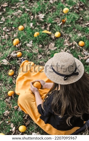 A pregnant Caucasian woman with long brown hair wearing a straw hat, sitting on the grass with lemons scattered around and holding one lemon in her hands