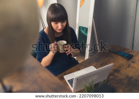 Young caucasian brunette woman with bangs wearing a dress sitting by a desk working on laptop holding a mug. Indoor shot. High quality photo