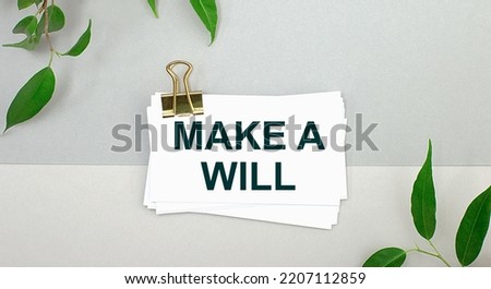 On a gray background - green leaves of the plant and a white card under a gold clip with the text MAKE A WILL. Minimalist.