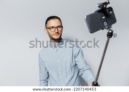 Studio portrait of young smiling man taking selfie photo via smartphone and stick. Light grey background. Wearing eyeglasses and blue t-shirt.