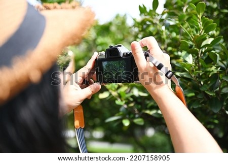 A female tourist wearing a brown straw hat uses a digital camera to photograph the beauty of the green trees in the park. Woman holding a small camera photographing leaves on blurred nature background