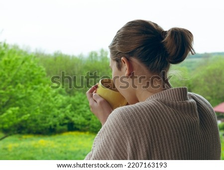 a woman in cool weather stands on the balcony and drinks coffee from a yellow cup looking at the courtyard