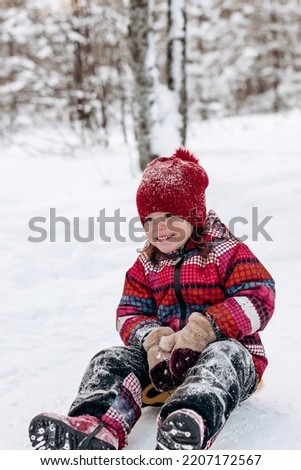 Happy little caucasian girl in a red hat and jumpsuit rides a saucer in the winter park.Beautiful trees are covered with white snow.Winter fun,active lifestyle concept.