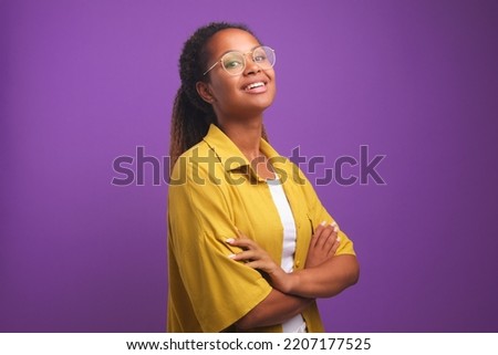 Young confident positive African American woman proudly posing with arms crossed in front of chest and raising chin looks at screen dressed in yellow shirt stands on purple studio background
