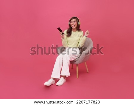 Happy young Asian teen woman smiling listening music in headphones and sitting on chair isolate on pink background.