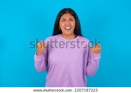 Portrait of Young latin woman wearing purple sweatshirt over blue background looks with excitement at camera, keeps hands raised over head, notices something unexpected reacts on sudden news.