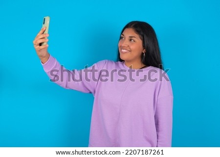 Isolated shot of pleased cheerful Young latin woman wearing purple sweatshirt over blue background , makes selfie with mobile phone. People, technology and leisure concept