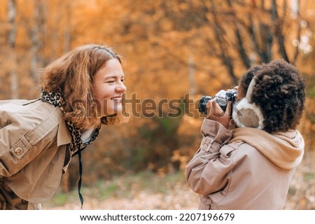 African-American daughter takes pictures of her caucasian mother on camera in an autumn park.Diversity,autumn concept.Side view,selective focus.