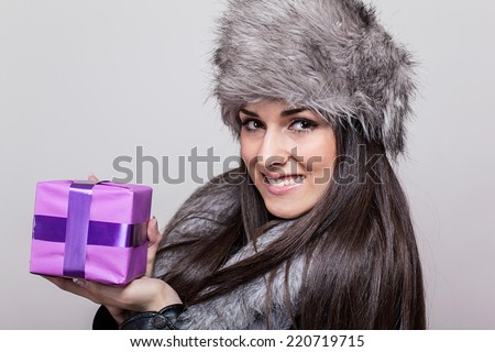 Beautiful young woman in fur hat holding present. Studio shot over gray background. 