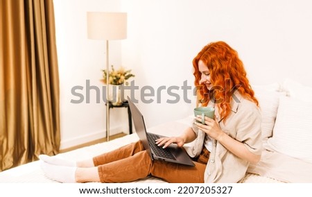 Smiling teen girl school student with orange hair use laptop computer sit on bed distance elearning online learning course prepare for exam search remote online course at home.