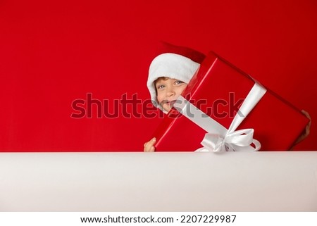 Happy Caucasian child in red hat holds in his hands large box with gift on red background. Smiling boy with Christmas gift box