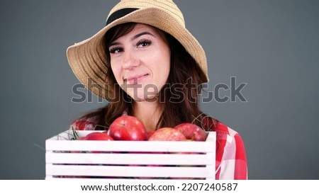 portrait of beautiful smiling female farmer in plaid shirt, gloves and hat holding white wooden box with red ripe organic apples, on gray background, in studio,. High quality photo