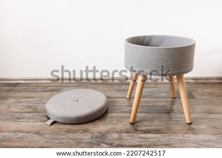 Trendy stool with and build-in storage space. Side view, white wall, copy space. Modern mulrifunctiional chair with wooden legs and removable lid. Round gray linen pouf Royalty-Free Stock Photo #2207242517